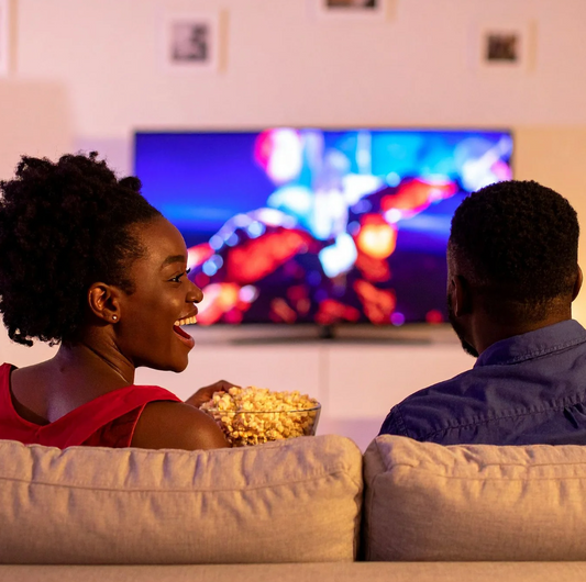 Two people sitting on a couch watching TV with popcorn, smiling and enjoying each other's company.